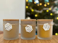 Three glass candles with wooden lids on a wooden surface, with a blurred Christmas tree in the background.